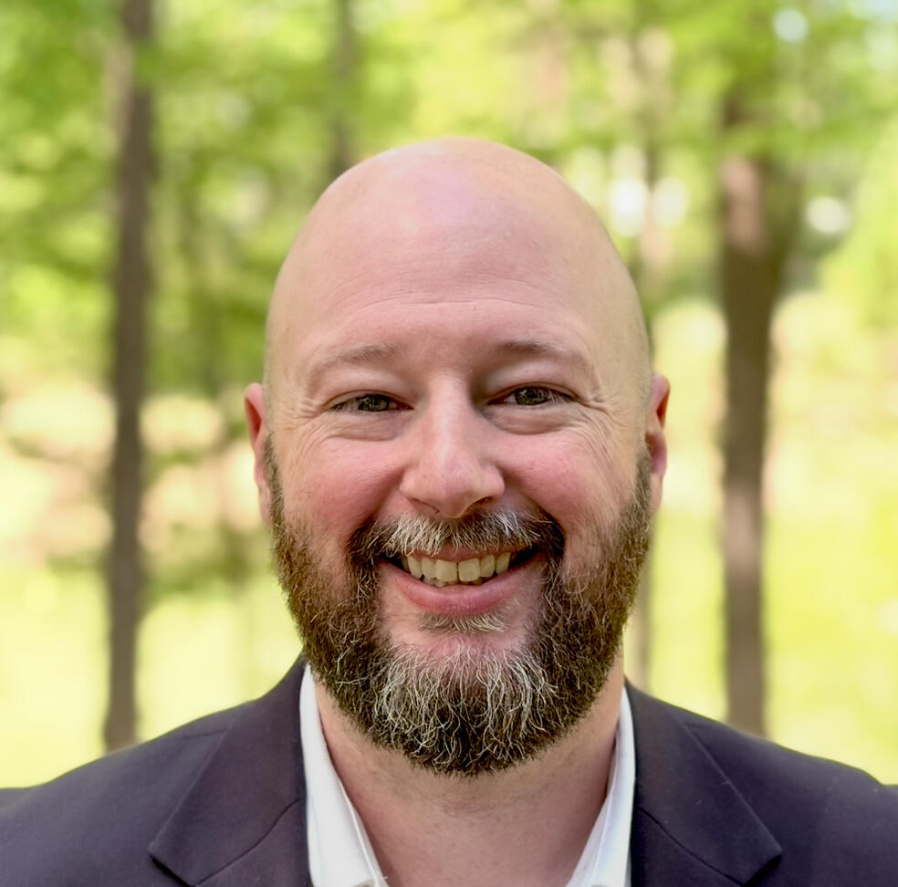 Shayne Martin with trees in the background, beard and bald, and middle aged with the look of experience.